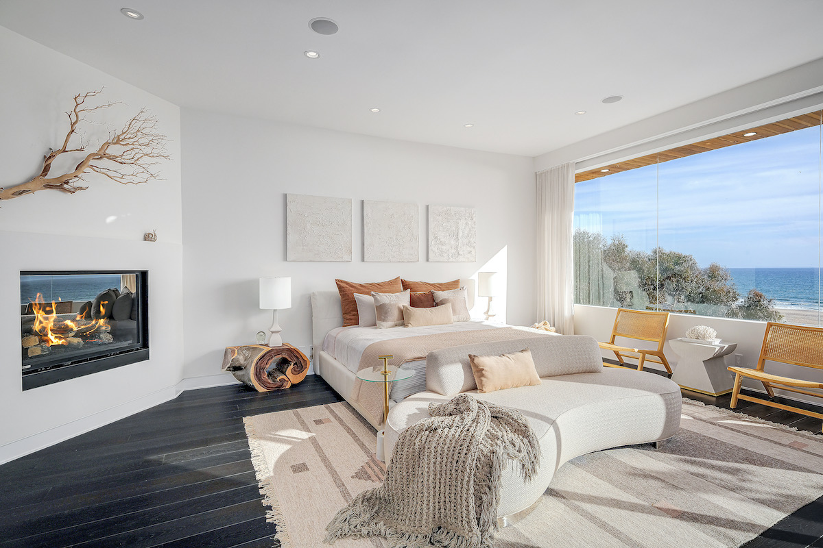 master bedroom with white walls and linens, a fireplace, 2 wooden chairs and a large window with ocean views