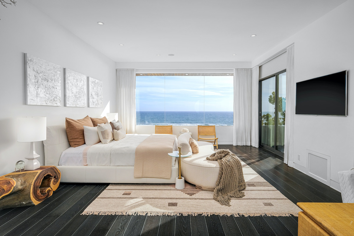 bedroom with white walls and linens, 2 wooden chairs, a tv on the wall, and large window with ocean views and a sliding door to the patio