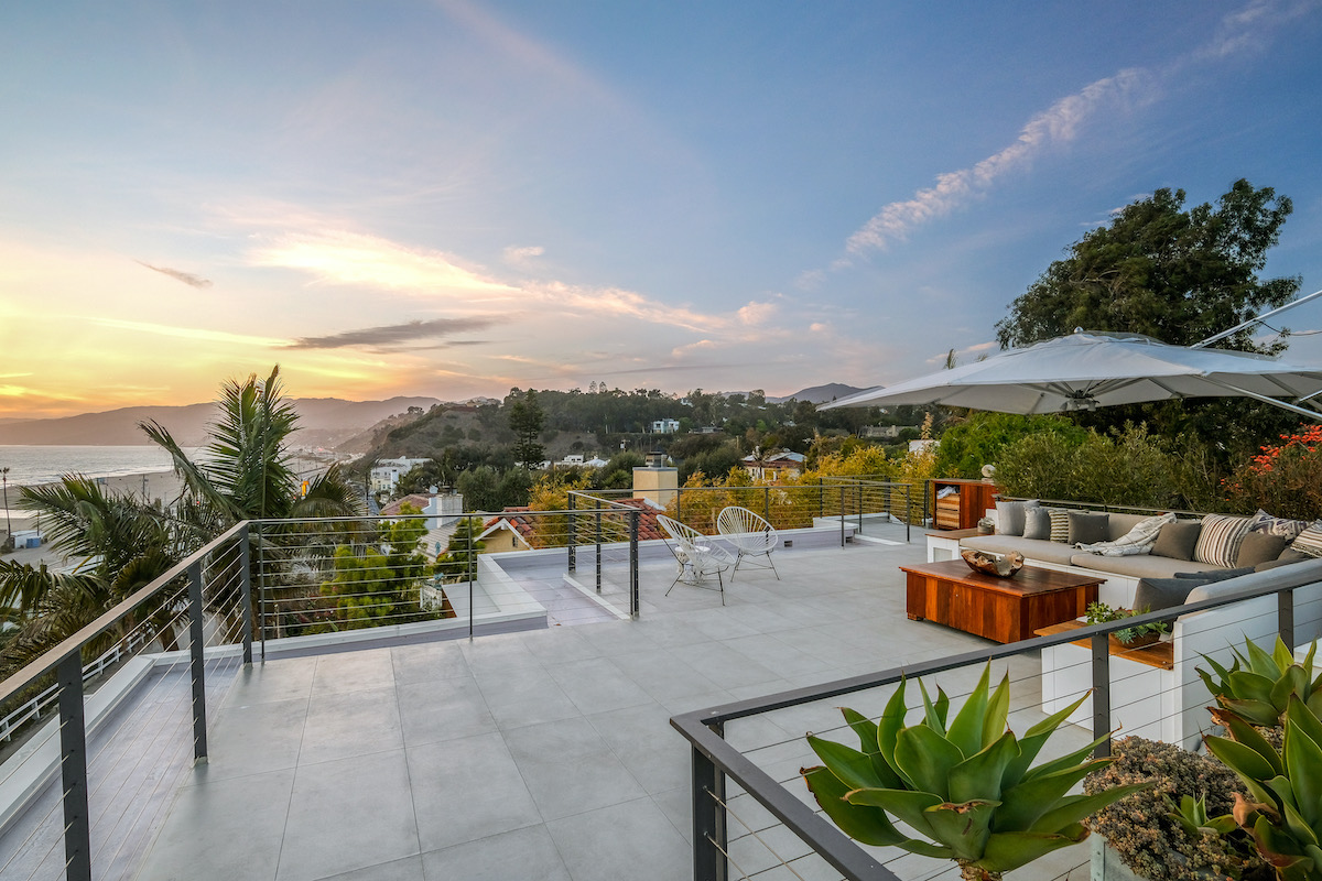 rooftop deck with a couch, wooden table, chairs, and umbrella looking at oceanfront views at sunset