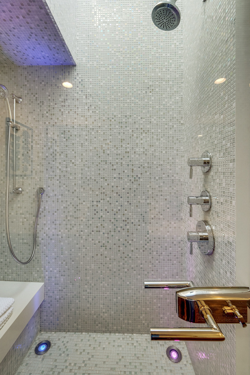 luxury shower with grey tile walls, two shower heads, and a shower seat